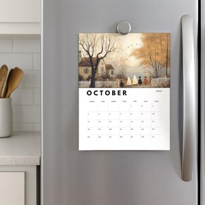 May include: A magnetic wall calendar for October 2027 featuring an illustration of a house, trees, and figures in costumes. The calendar has a white background with black text and a grid for dates. It is attached to a stainless steel surface.