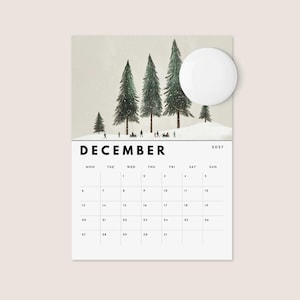 May include: A December 2027 wall calendar page featuring a watercolor illustration of snow-covered pine trees and small figures in a winter landscape. The calendar grid is printed in black on a white background with a circular white magnet overlay.