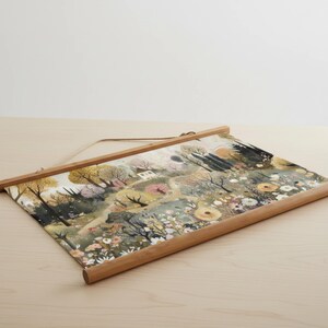 May include: A landscape art print featuring a watercolor painting of a field with trees, flowers, and a small house. The print is held by a wooden frame with a hanging string. The color palette includes greens, yellows, and pinks.