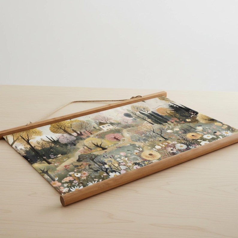 May include: A landscape art print featuring a watercolor painting of a field with trees, flowers, and a small house. The print is held by a wooden frame with a hanging string. The color palette includes greens, yellows, and pinks.