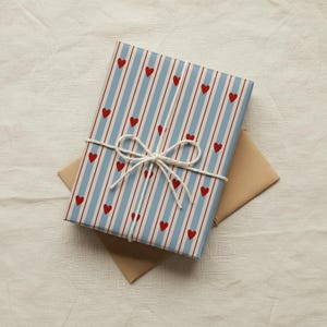 May include: A wrapped gift with blue and white striped paper, adorned with red hearts. The package is tied with white twine, forming a bow. A brown envelope rests beneath the gift, all on a white fabric surface.