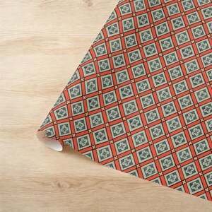 May include: A roll of wrapping paper featuring a repeating geometric grid pattern. The design consists of light green squares, each containing a small four-petaled flower, separated by bold orange bands and small black cross accents.