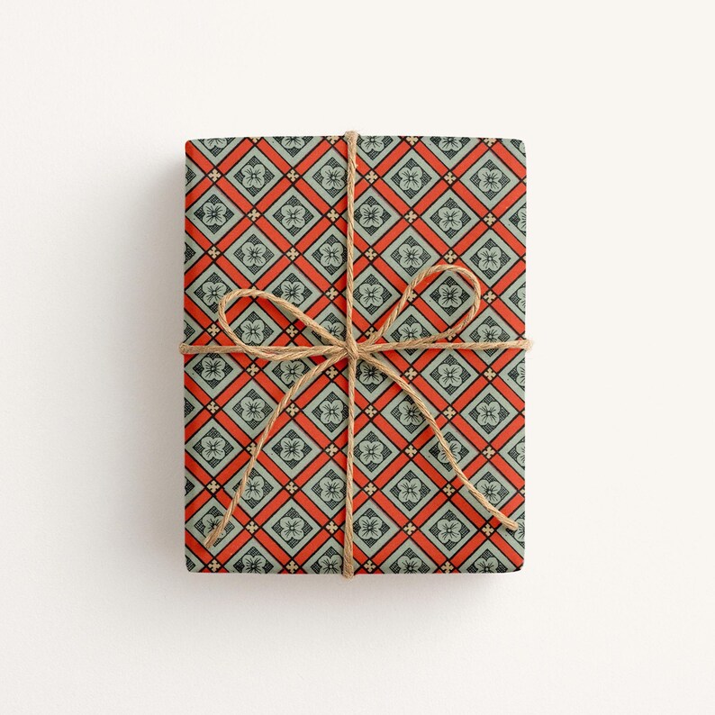 May include: A rectangular gift wrapped in paper featuring a repeating geometric pattern of orange diamonds and light blue squares, each containing a small four-petaled flower motif. The package is tied with a thin, natural brown jute twine bow.