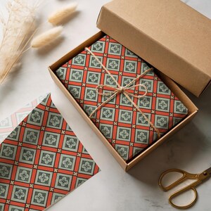May include: A rectangular cardboard box containing a gift wrapped in paper with a repeating geometric pattern of orange squares and light green floral motifs, tied with thin brown twine. A matching sheet of wrapping paper lies nearby.