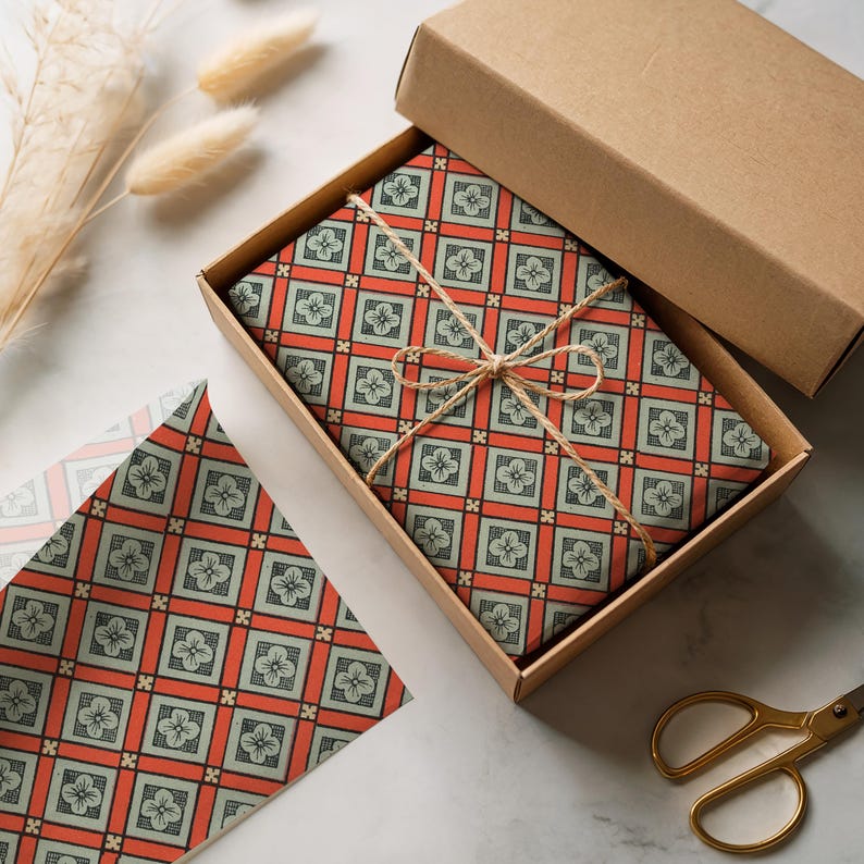 May include: A rectangular cardboard box containing a gift wrapped in paper with a repeating geometric pattern of orange squares and light green floral motifs, tied with thin brown twine. A matching sheet of wrapping paper lies nearby.