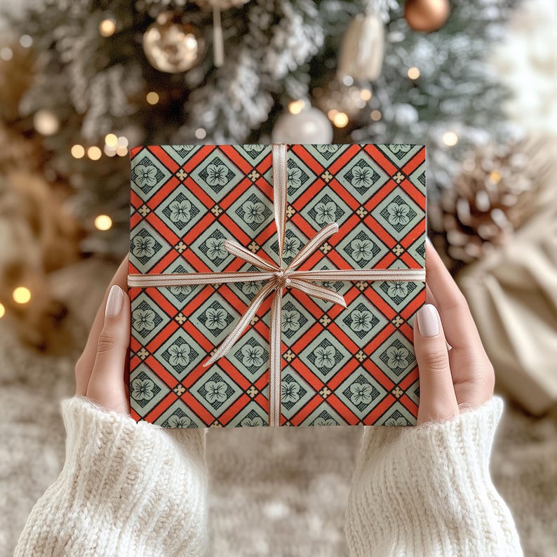 May include: A square gift box wrapped in paper featuring a repeating geometric pattern of orange diamonds and light green squares with small floral motifs, tied with a thin, light-coloured ribbon centred in a bow across the front surface.