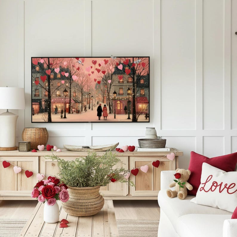 May include: A living room scene with a large framed artwork depicting a romantic street scene with heart-shaped balloons. A wooden cabinet holds a woven basket, flowers, and heart garlands. A white sofa has red pillows and a teddy bear. The word "Love" is embroidered on a pillow.