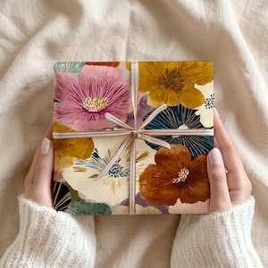 May include: A square gift box wrapped in paper featuring a vibrant floral pattern with large blooms in shades of pink, mustard yellow, deep brown, and cream, tied with a thin, light-colored ribbon that crosses in the center of the package.