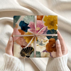 May include: A wrapped gift box featuring a floral pattern in shades of pink, yellow, brown, and blue. The gift is tied with a striped ribbon. The box is held by a person wearing a white sweater.