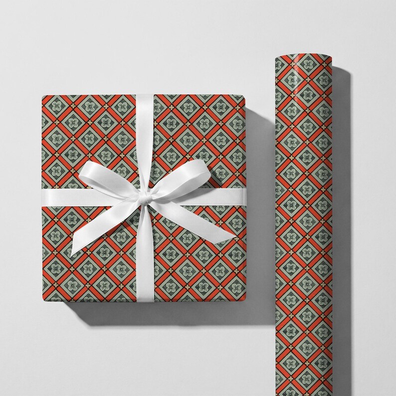 May include: A square gift box wrapped in paper with a repeating geometric diamond pattern in orange, dark green, and cream, tied with a white satin ribbon bow, alongside a matching roll of the same patterned wrapping paper standing vertically.