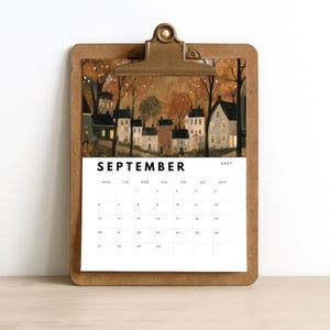 May include: A brown wooden clipboard holding a September 2027 calendar page. The top section features an illustration of a small village with houses, trees, and warm glowing lights at dusk. The calendar grid is printed in black on a white background.