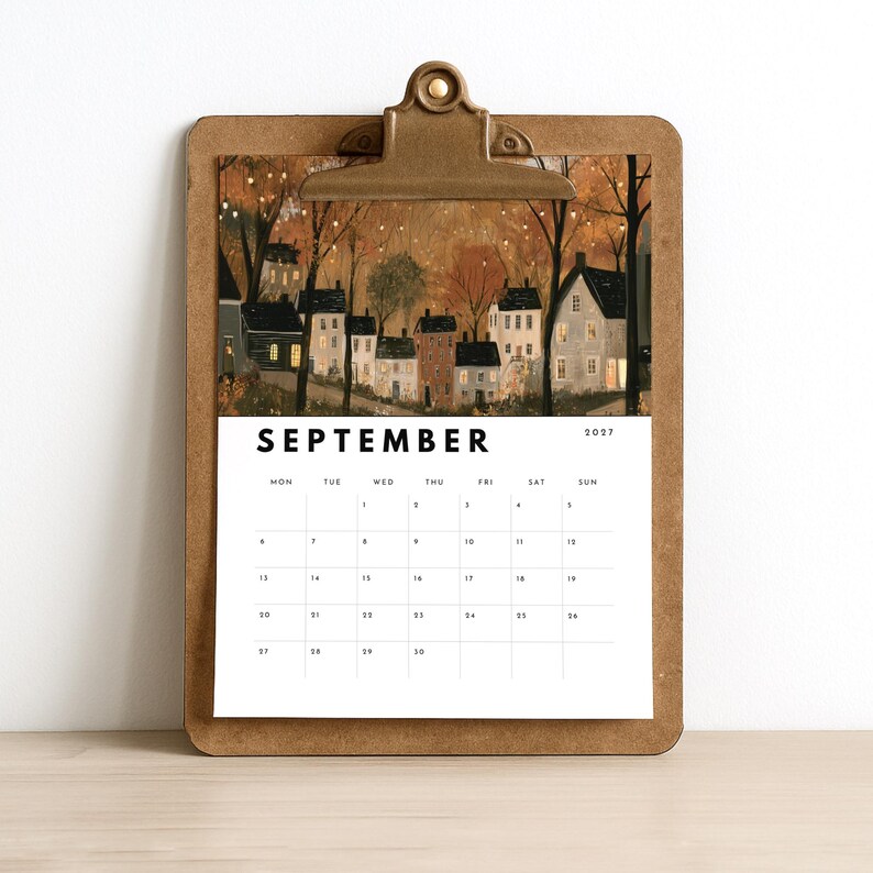 May include: A brown wooden clipboard holding a September 2027 calendar page. The top section features an illustration of a small village with houses, trees, and warm glowing lights at dusk. The calendar grid is printed in black on a white background.