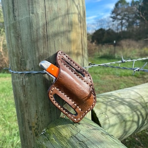 May include: A brown leather knife sheath with a stitched design and a small pocket knife with an orange handle inside.