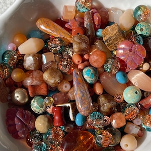 May include: A collection of assorted loose beads in a white bowl, featuring a variety of shapes, sizes, and textures. The beads include translucent glass, opaque stone, metallic finishes, and iridescent surfaces in shades of orange, blue, and brown.