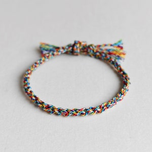 Puede incluir: Un brazalete trenzado multicolor con cierre anudado y borlas en cada extremo.