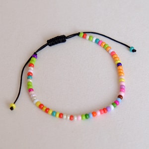 以下が含まれることがあります： 黒い紐とスライド式の結び目留め具が付いたカラフルなビーズブレスレット。ビーズは赤、オレンジ、黄色、緑、青、紫、白など、さまざまな色です。