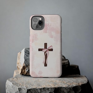 Puede incluir: Funda de teléfono con una cruz marrón y una cinta rosa, sobre un fondo de acuarela rosa pálido. La funda está diseñada para un smartphone y tiene un acabado brillante. El diseño tiene un tema delicado y religioso.