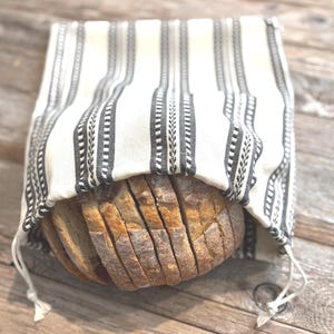May include: A rectangular fabric bread bag featuring a cream-colored base with vertical black stripes and intricate woven patterns. The bag is open at the top, revealing a sliced loaf of crusty, golden-brown sourdough bread inside the pouch.