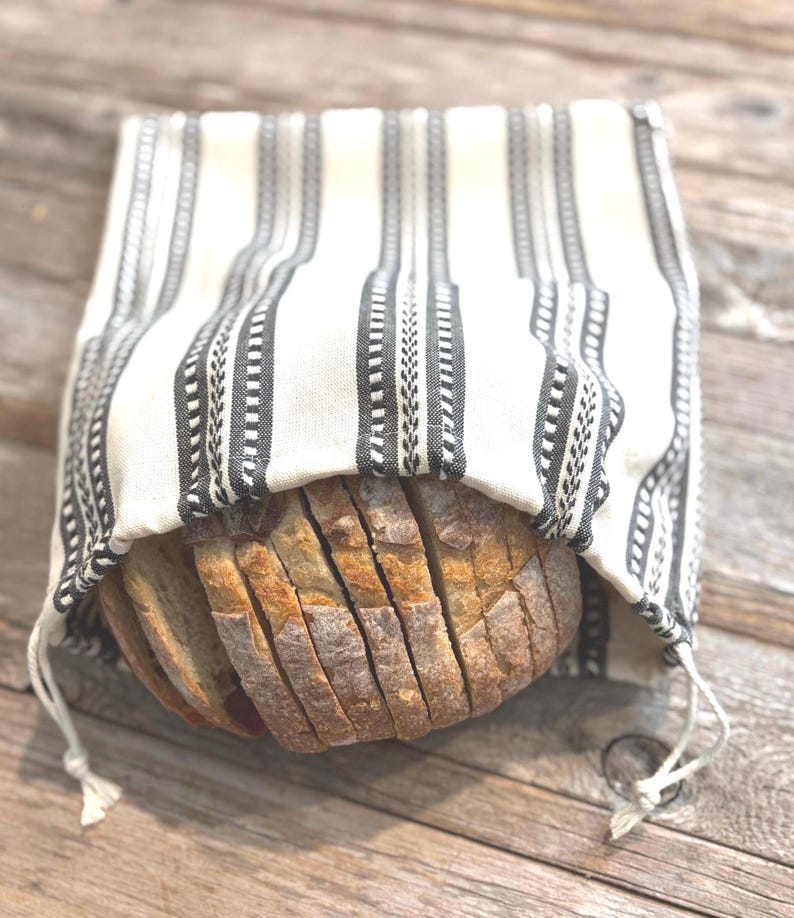 May include: A rectangular fabric bread bag featuring a cream-colored base with vertical black stripes and intricate woven patterns. The bag is open at the top, revealing a sliced loaf of crusty, golden-brown sourdough bread inside the pouch.