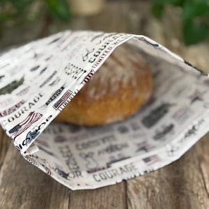 May include: A white bread bag with a patriotic print, holding a round loaf of bread. The bag features American flags, military symbols, and words like "Courage" and "Liberty". The bread is golden brown.