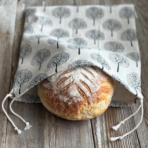May include: A round loaf of bread peeks out from a beige bread bag. The bag is patterned with black tree silhouettes. The bread has a scored design on top. The bag has a drawstring closure with white cords.