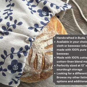 May include: A beige fabric bread bag with a dark blue floral vine pattern containing a round loaf of sourdough bread. A dark gray text box lists product details including dimensions of 13 inches by 13.5 inches and material options.