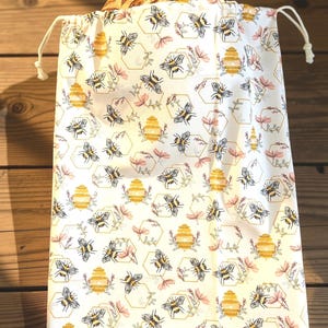 May include: A white fabric drawstring bag featuring a repeating pattern of yellow beehives, black and yellow bees, and small pink floral sprigs enclosed in thin gold hexagonal outlines. The bag is filled with a round loaf of crusty bread.