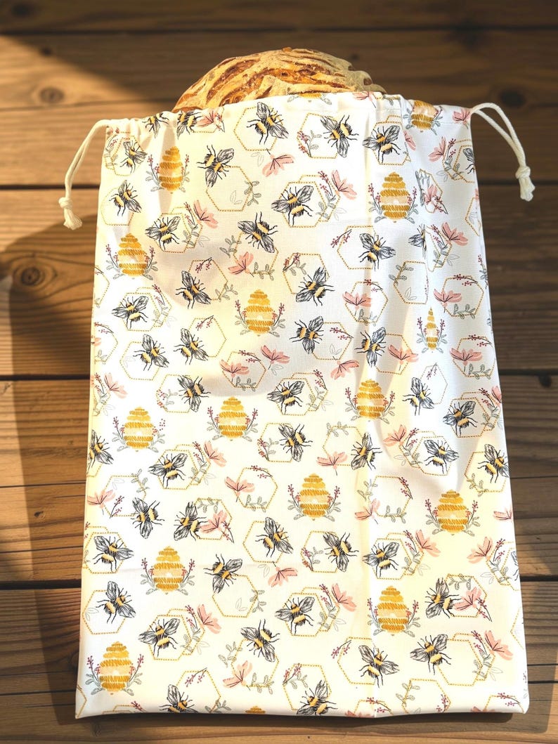 May include: A white fabric drawstring bag featuring a repeating pattern of yellow beehives, black and yellow bees, and small pink floral sprigs enclosed in thin gold hexagonal outlines. The bag is filled with a round loaf of crusty bread.