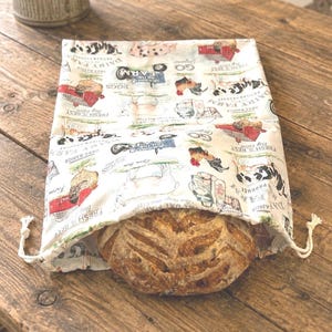 May include: A cream-colored fabric bread bag with a drawstring closure, featuring a repeating farm-themed print with red barns, cows, chickens, tractors, and text including "Dairy Farm" and "Fresh is Best," containing a round loaf of crusty bread.