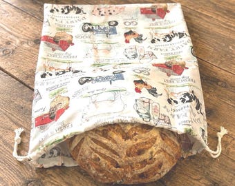 May include: A cream-colored fabric bread bag with a drawstring closure, featuring a repeating farm-themed print with red barns, cows, chickens, tractors, and text including "Dairy Farm" and "Fresh is Best," containing a round loaf of crusty bread.