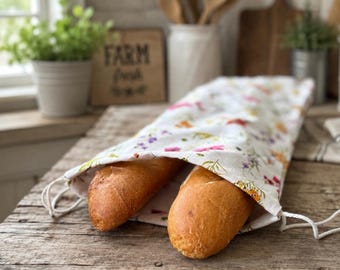May include: A white fabric bread bag featuring a colorful floral pattern with small pink, yellow, and purple flowers and green leaves. The bag has a drawstring closure and contains two golden-brown baguettes protruding from the open end.