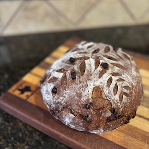 May include: A round loaf of artisan bread with a leaf pattern cut into the crust, dusted with flour, and studded with dark inclusions. The bread rests on a wooden cutting board with a dark brown border.