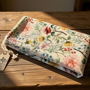 May include: A folded rectangular fabric pouch featuring a cream background with a colorful floral pattern of pink, yellow, and blue flowers and green leaves. A white rope drawstring is attached to a tan cardboard tag labeled BUDAFUL CREATIONS.