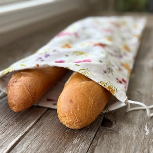 May include: A white bread bag with a floral pattern holds two golden-brown baguettes. The bag has a drawstring closure. The bag and bread are on a weathered wooden surface.