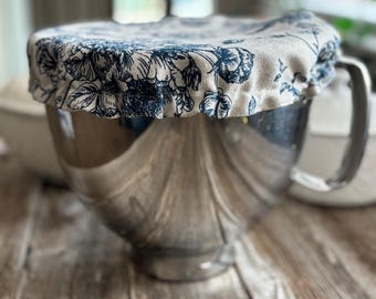 May include: A stainless steel mixing bowl with a handle, covered by a circular fabric bowl cover. The cover features a beige background with a dark blue floral pattern and an elasticized edge that fits snugly around the rim of the bowl.