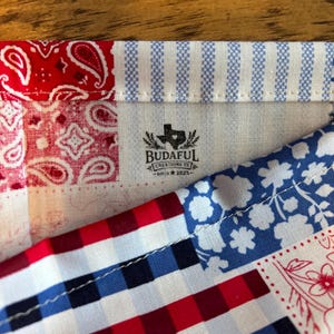 May include: A fabric patchwork textile featuring red paisley, blue and white stripes, blue floral patterns, and red, white, and blue gingham checks. A central logo reads BUDAFUL CREATIONS TX SINCE 2025 with a Texas state silhouette icon.