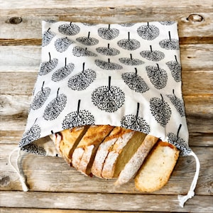 May include: A white fabric drawstring bag featuring a repeating black tree pattern. The bag is partially open, revealing several thick, golden-brown slices of crusty bread inside. The fabric has a soft texture and white cord drawstrings.