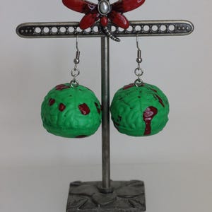 May include: A pair of unique earrings featuring green, brain-shaped beads with red accents, suspended from a decorative metal stand. A red dragonfly tops the stand. The earrings are a quirky, artistic accessory.