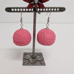 May include: A pair of pink brain-shaped earrings hanging from a decorative metal display stand. The stand features a dragonfly accent and a floral base. The earrings are a vibrant pink color and have a detailed brain texture.