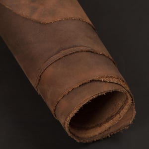 May include: A rolled-up length of distressed brown leather. The leather has a textured surface with visible grain and a worn appearance. The edges are raw and unfinished, giving a rustic look. This material is likely used for crafting items such as bags or belts.
