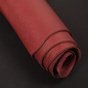May include: A roll of red leather, tightly curled. The leather has a textured surface and the edges of the material are visible. The background is a plain black.