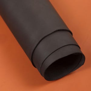May include: A rolled sheet of dark brown leather, revealing its smooth texture and layers. The leather is set against an orange background, highlighting its rich colour and potential for crafting projects.