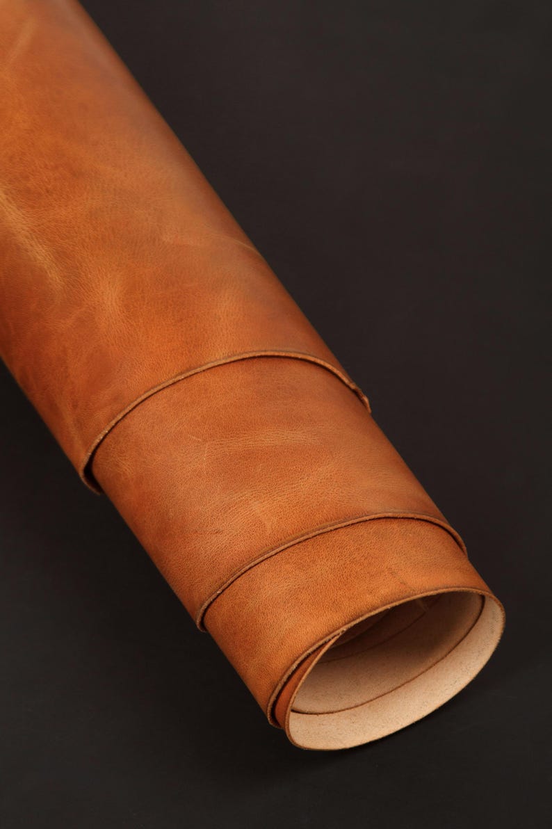 May include: A rolled-up piece of tan leather, showcasing its natural grain and texture. The leather is a warm, light brown color, with visible creases and subtle variations in tone. The edges are neatly rolled, revealing a lighter interior. The background is a solid black.