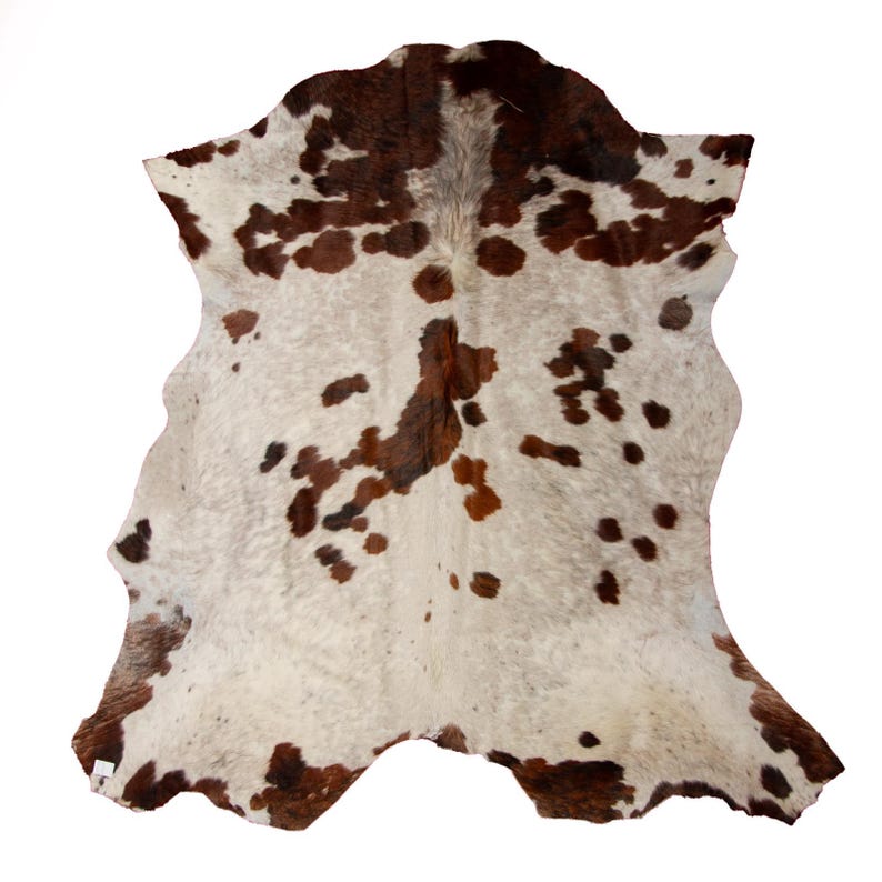 May include: A white cowhide rug with brown and reddish-brown spots. The rug is folded in half and has a natural, rustic look.