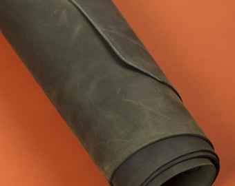 May include: Rolled-up, dark olive-green leather material. The leather has a distressed, textured appearance with visible grain and slight color variations. The edges of the roll are visible, revealing the thickness of the material. The background is a solid orange.