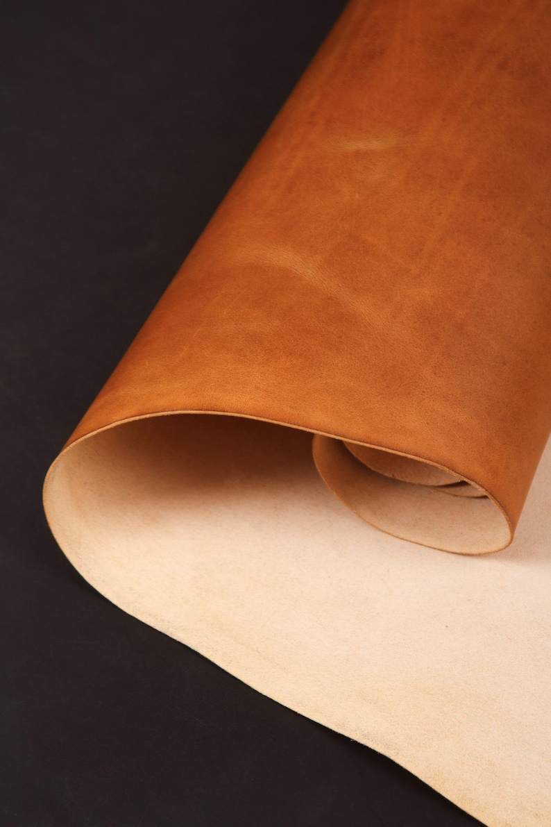 May include: A rolled sheet of tan leather with a smooth, textured surface. The leather is a warm, earthy tone, with a lighter, natural underside. The background is a dark, solid color.