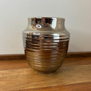 Puede incluir: Un jarrón metálico con un cuerpo ancho y redondeado y un cuello estrecho. El jarrón tiene un acabado plateado y bronce con detalles horizontales acanalados alrededor del centro. Se encuentra sobre una bandeja de madera.