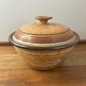 Puede incluir: Un cuenco de cerámica con tapa. El cuenco y la tapa son de color marrón claro con detalles marrones y negros. La tapa tiene un pequeño asa en la parte superior. El cuenco está sobre una superficie de madera.
