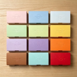 Puede incluir: Una colección de doce cajas rectangulares de cartón dispuestas en una cuadrícula, cada una con el texto en relieve naturaloop. Las cajas son de color rosa, azul claro, blanco, verde claro, verde lima, amarillo, lavanda, morado, naranja, marrón, cerceta y rojo.