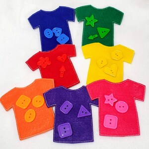 May include: Six felt t-shirts in different colors, each with a different button and shape on them. The colors are blue, green, red, yellow, orange, and purple.